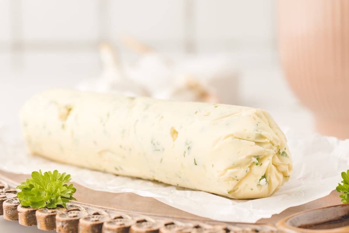 A log of garlic compound butter wrapped in parchment paper sits on a wooden board, with a sprig of parsley nearby.
