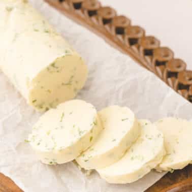 A log of garlic compound butter with several round slices cut, displayed on parchment paper atop a wooden tray.