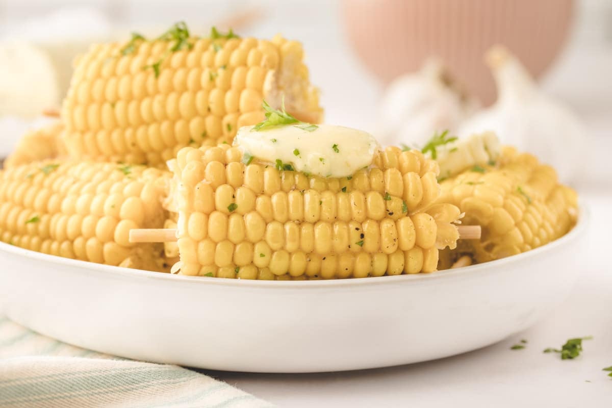 A plate of corn on the cob with a pat of garlic compound butter and chopped herbs on top, served on wooden skewers.