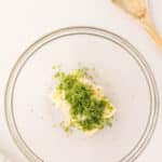 A glass bowl containing butter and chopped green herbs next to a wooden spoon on a white surface.