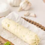 A log of garlic compound butter rests on parchment paper atop a wooden tray, with garlic bulbs and parsley in the background.