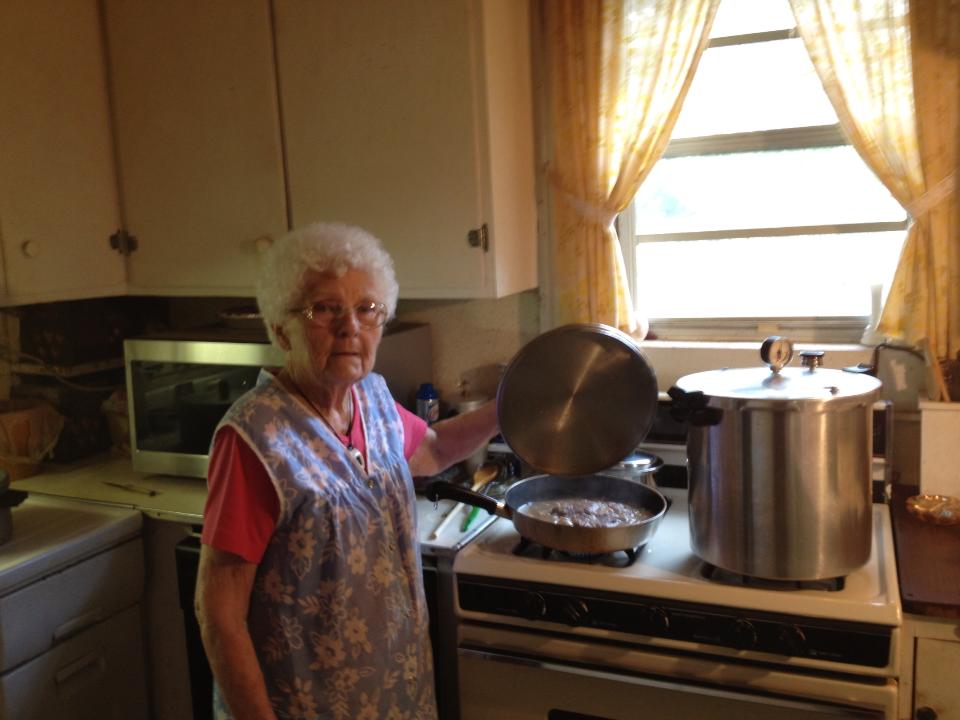 An older woman stands in a kitchen, lifting the lid off a pot on the stove next to a large pressure cooker.