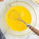 A glass bowl with beaten eggs and a whisk, on a marble countertop next to eggshells, a baking dish, and a gray cloth.
