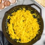 Scrambled eggs cooked in a black cast iron skillet on a marble countertop, with empty eggshells and a baking dish nearby.