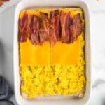 A rectangular baking dish contains a layer of scrambled eggs, topped with cheddar cheese slices and strips of cooked bacon.