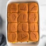 A white baking dish filled with twelve baked slider sandwiches, arranged in a 3 by 4 grid, on a marble countertop.