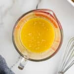 A glass measuring cup filled with yellow liquid, speckled with sesame seeds, sits on a marble surface next to a metal whisk.