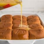 A glass measuring cup pours melted butter over a batch of dinner rolls in a white baking dish.