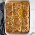 A baking dish filled with twelve slider sandwiches topped with a grainy mustard and butter mixture, sitting on a marble surface next to a dark cloth napkin.
