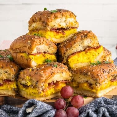 A stack of breakfast sliders with scrambled eggs, bacon, and cheese on poppy seed buns, arranged on a wooden board with grapes and berries in the background.
