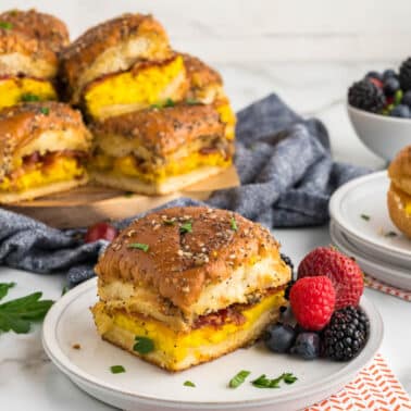 Hawaiian roll breakfast slider with eggs, sausage, and cheese on buns are served on plates with fresh mixed berries, with more sliders and berries in the background.