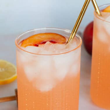 Two tall glasses of iced peach lemonade, each garnished with a peach slice and gold straw, with lemon slices and a peach in the background—perfect for enjoying flavorful lemonades and refreshing summer drinks.