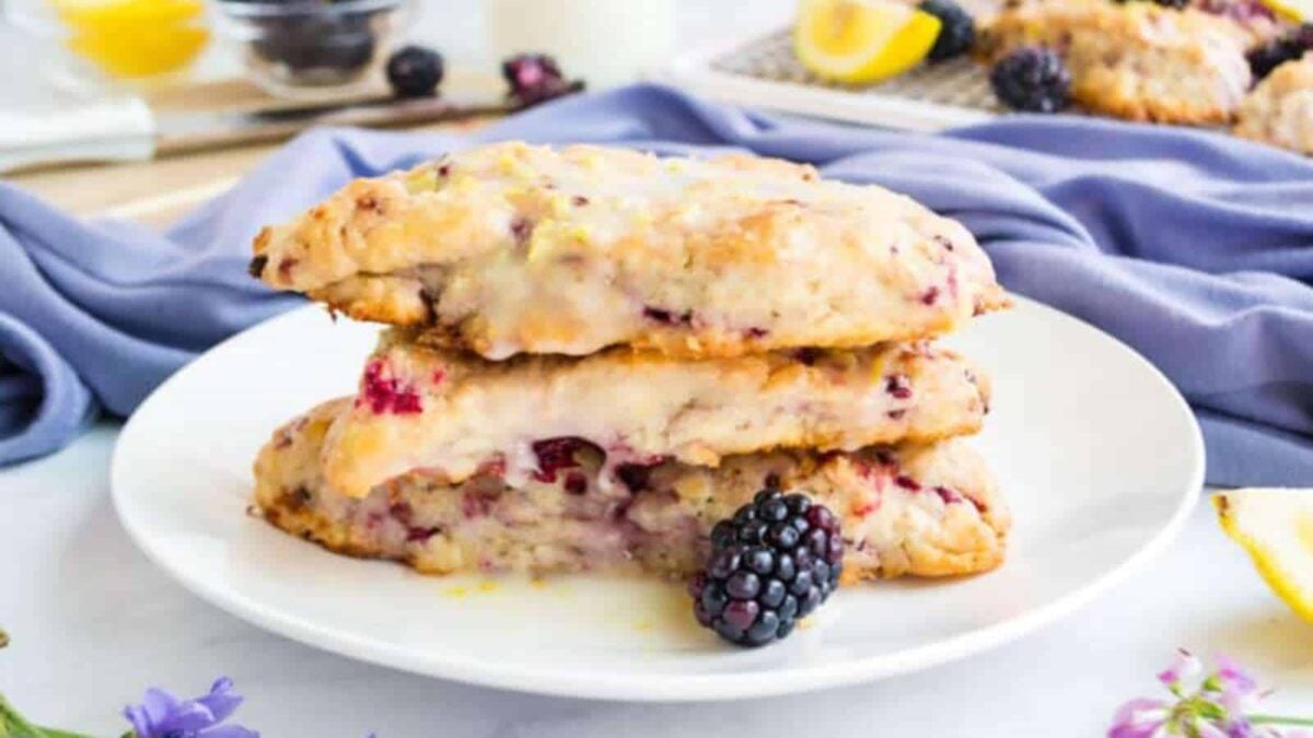 Two berry scones are stacked on a white plate, with blackberries blending into the dough. A blackberry rests in front, and other scones and ingredients are blurred in the background—perfect for those who love blackberry recipes and baking.