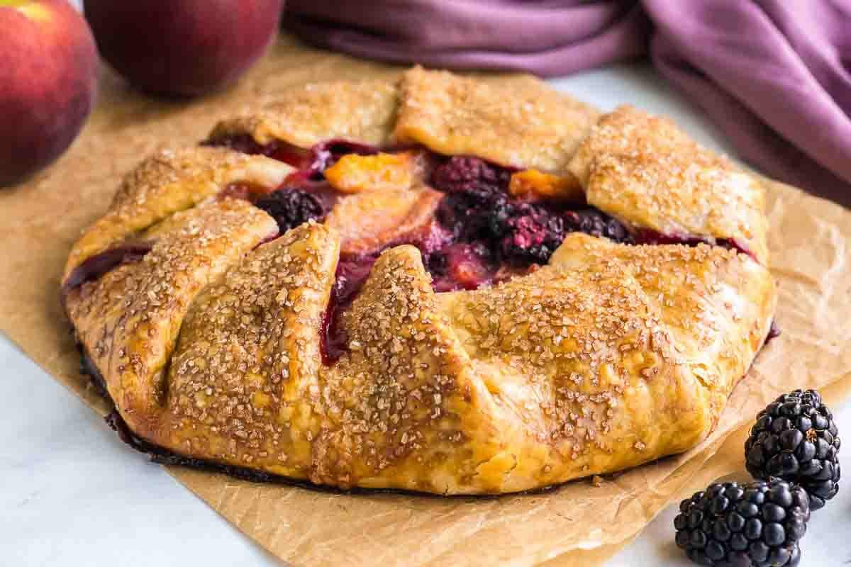 A rustic fruit galette with a golden, sugared crust filled with peaches and blackberries sits on parchment paper, perfect for anyone who loves baking or experimenting with new blackberry recipes. Fresh peaches and blackberries rest nearby.