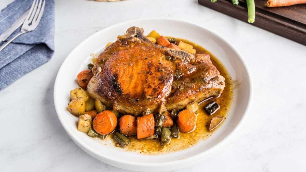 A cooked pork chop with brown gravy is served on a white plate with roasted carrots, potatoes, and green beans—a classic favorite among pork chop recipes.