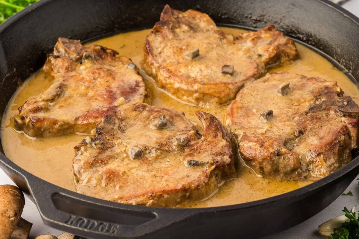 Four cooked pork chops in a creamy mushroom sauce are shown in a black cast iron skillet, making this one of the coziest pork chop recipes to try.