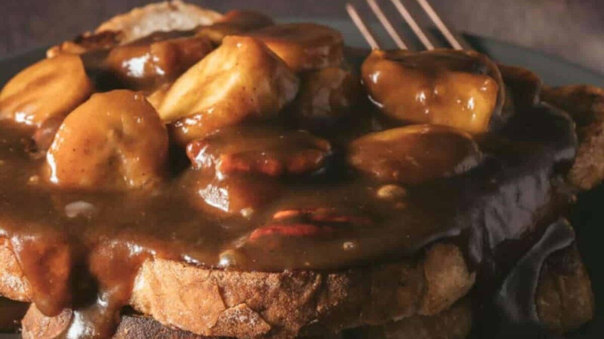 Slices of bread topped with thick, brown gravy containing pieces of sausage and banana make for a unique twist on sweet breakfast ideas, served on a dark plate with a fork in the background.
