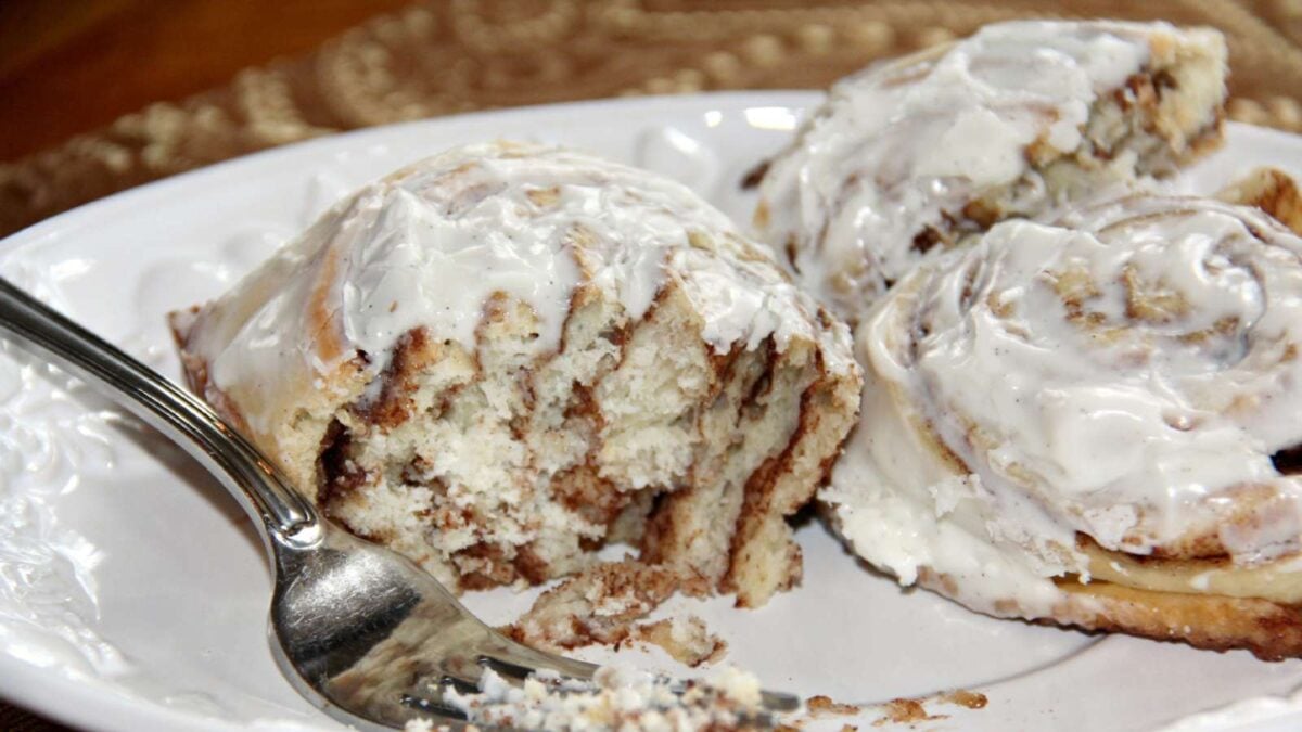 Two cinnamon rolls with white icing on a white plate, one partially eaten with a fork beside it—a delicious addition to your morning meals or sweet breakfast ideas.