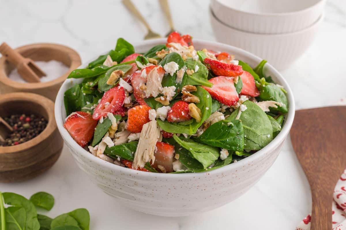 A bowl of spinach salad topped with sliced strawberries, crumbled cheese, chopped nuts, and shredded chicken sits on a white countertop with small bowls of salt and pepper nearby—perfect for those seeking easy spinach recipes.