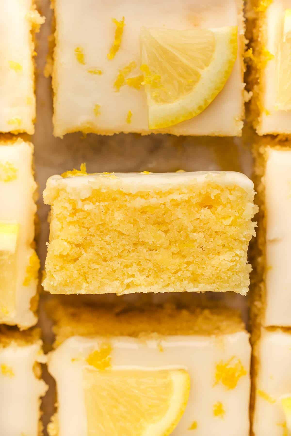 Close-up of lemon brownies topped with white icing and lemon zest, with one bar turned sideways to show the moist, yellow interior.