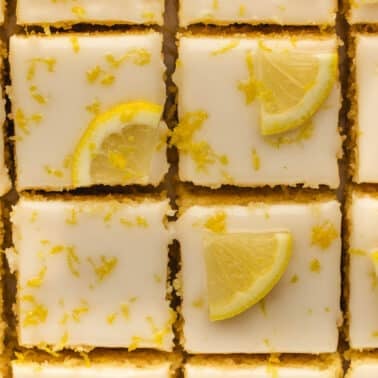 Rows of lemon brownies topped with white icing, lemon wedges, and lemon zest, arranged closely together in a grid pattern.