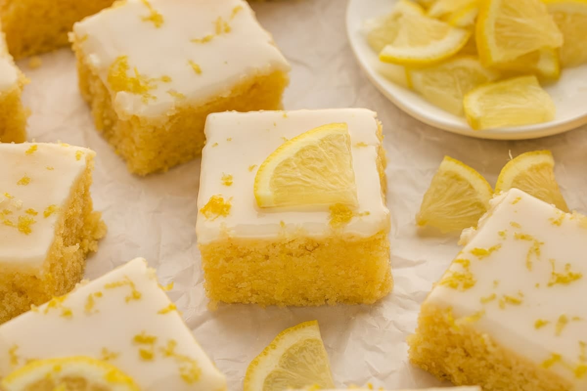 Lemon brownies with white icing, topped with lemon slices and zest, arranged on parchment paper with a plate of lemon slices in the background.