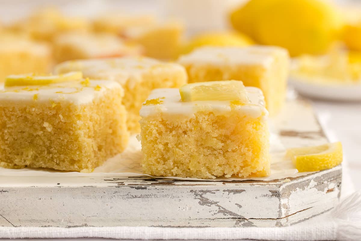 Lemon brownies with white icing and lemon slices on top, arranged on a rustic white wooden surface.