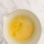 A mixing bowl containing a yellow, frothy liquid mixture sits on a white marble surface next to a white cloth.