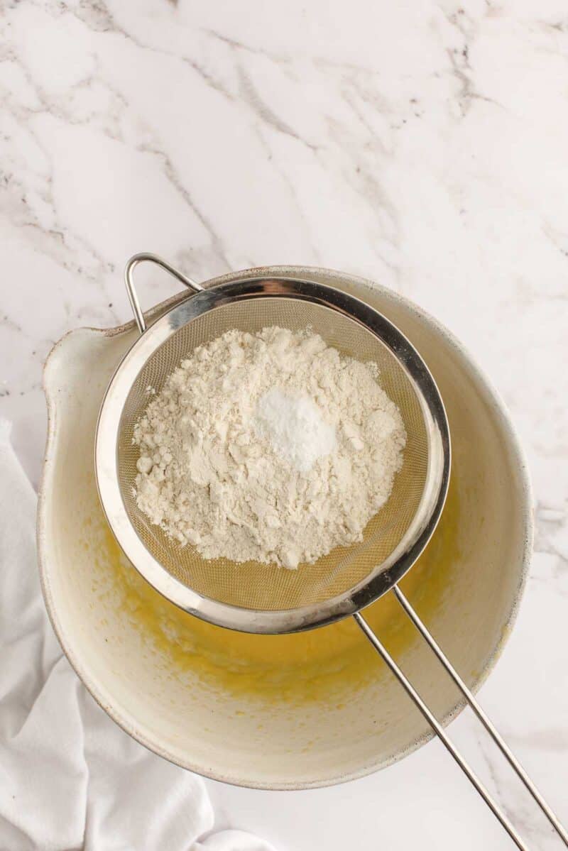 A metal sifter holding flour and baking powder is placed over a mixing bowl containing a yellow batter, on a marble countertop.