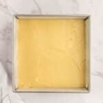 A square metal baking pan filled with smooth, unbaked yellow cake batter on a white marble surface.