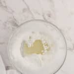 A glass bowl containing powdered sugar and a pool of liquid, set on a white marble surface with part of a white cloth visible.