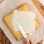 A hand spreads white icing onto a square sponge cake with a spatula, on parchment paper over a wooden board.