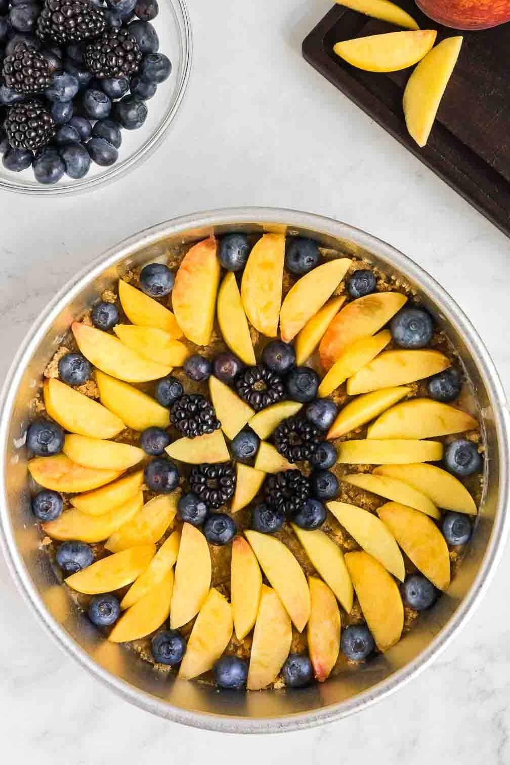 A round metal baking dish filled with a peach upside down cake sits on a white surface. A bowl of blueberries, blackberries, and a peach with slices are next to it.