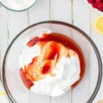 A glass bowl containing whipped cream and a swirl of raspberry sauce sits on a white wooden surface, surrounded by a bowl of whipped cream, lemon halves, raspberries, and a wooden spoon.