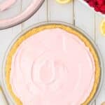 A raspberry lemonade pie with a pink creamy filling in a crumb crust sits on a white wooden table, surrounded by lemon slices and a bowl of raspberries.