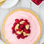 A raspberry lemonade pie topped with fresh raspberries and lemon slices on a glass plate, with plates, gold forks, and a bowl of raspberries nearby on a white table.