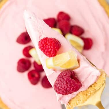 A slice of pink cheesecake topped with raspberries and lemon pieces is held above the rest of the cheesecake, which is garnished with fresh raspberries.