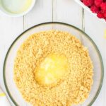 A glass bowl filled with graham cracker crumbs and melted butter, surrounded by a halved lemon, a bowl of raspberries, and a small dish of lemon juice on a white wooden surface.