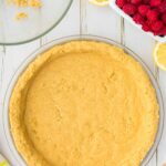 A pie dish with an unbaked crust, a glass bowl with crumbs, fresh raspberries in a dish, lemon slices, and a yellow striped towel on a white wooden surface.