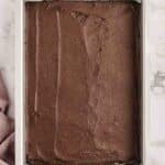 A rectangular baking pan filled with an even layer of unbaked chocolate batter sits on a marble surface next to a mauve cloth.