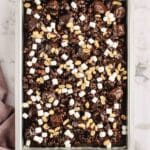 A baking pan filled with rocky road brownies topped with mini marshmallows and peanuts, set on a marble surface with a purple cloth nearby.
