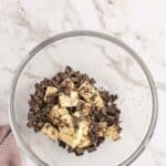 A glass bowl on a marble surface contains chunks of butter and chopped chocolate. A mauve cloth is partially visible beside the bowl.