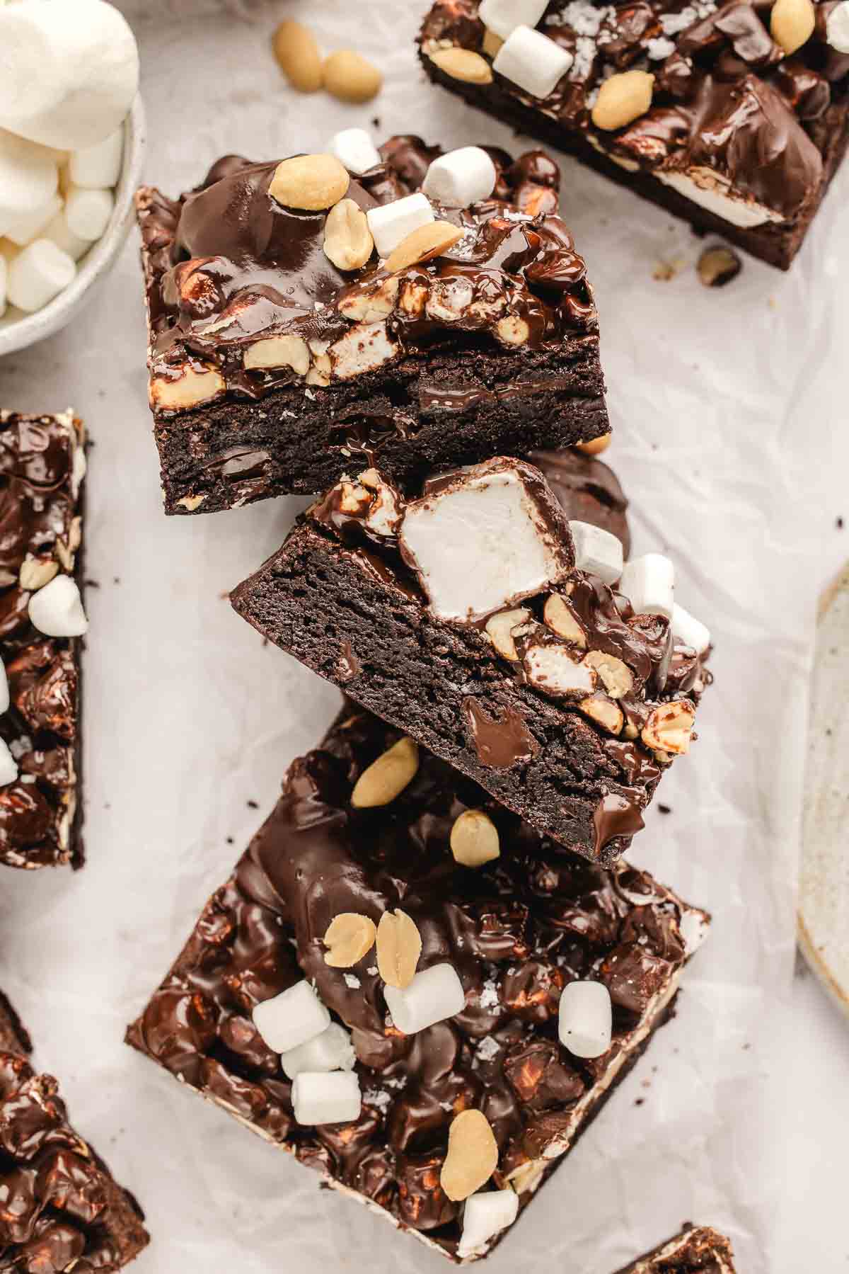 Three stacked rocky road brownies with marshmallows and peanuts on top, surrounded by more brownies and a small bowl of marshmallows on a white surface.