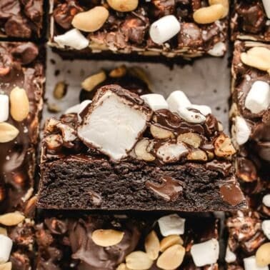 Close-up of a rocky road brownie topped with peanuts, mini marshmallows, and a chocolate coating, surrounded by similar brownies.