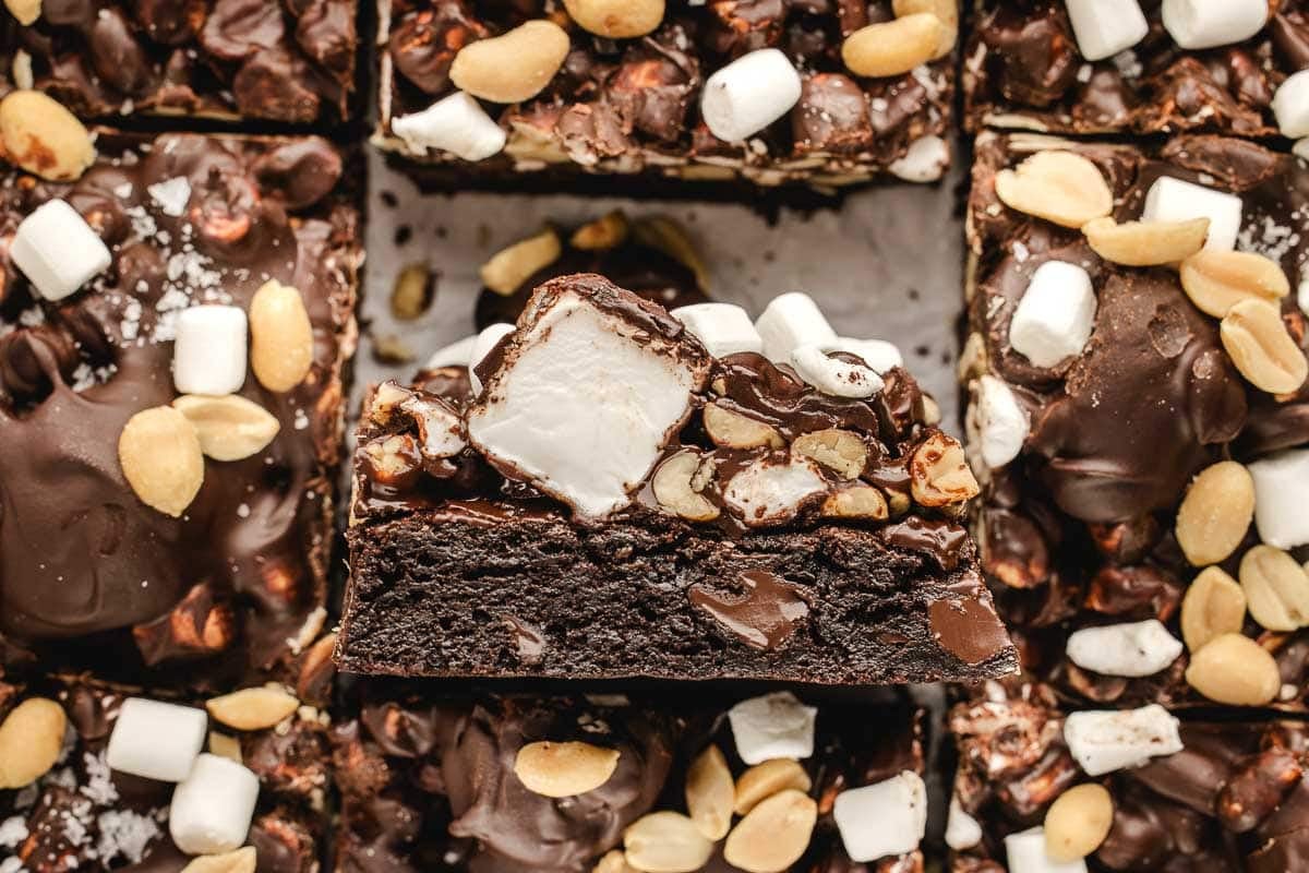 Close-up of a rocky road brownie topped with peanuts, mini marshmallows, and a chocolate coating, surrounded by similar brownies.