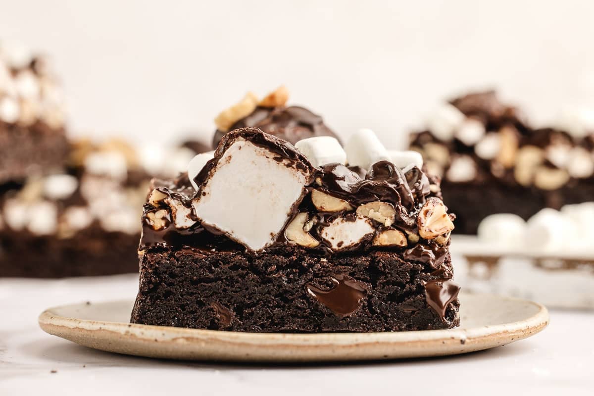 A slice of rocky road brownie topped with marshmallows, chopped nuts, and a drizzle of chocolate frosting, served on a beige plate.