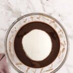A glass bowl containing chocolate batter with a mound of white sugar on top sits on a marble countertop next to a mauve cloth.