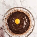 A glass bowl containing chocolate batter with a raw egg in the center, placed on a marble countertop next to a mauve cloth.