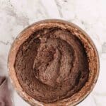 A glass bowl filled with chocolate cake batter sits on a white marble surface next to a light purple cloth.
