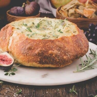 A round bread bowl filled with dip is garnished with herbs and placed on a marble serving board, surrounded by fresh figs, grapes, crackers, and rosemary.